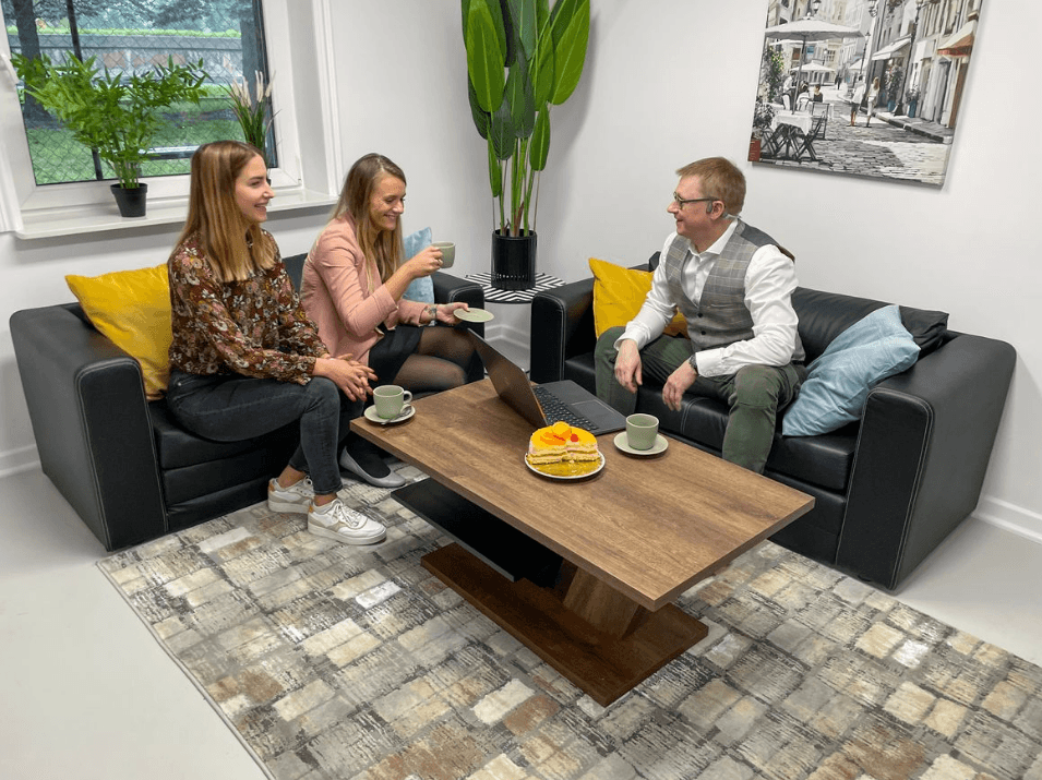 Three people enjoying a casual meeting with coffee and cake in a modern living room with stylish decor and a cityscape wall art