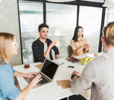 Four colleagues in a casual meeting at a modern office, with two men and two women around a table, discussing and using a laptop and tablet, with a bowl of apples in the center of the table.